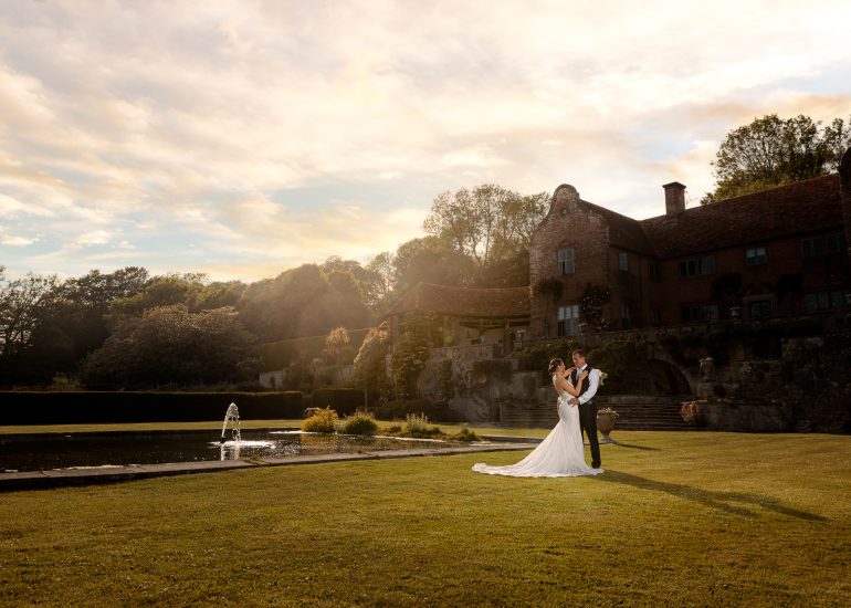 Bride and groom embracing by a pond in front of a historic building at sunset, captured by an Essex wedding photographer.