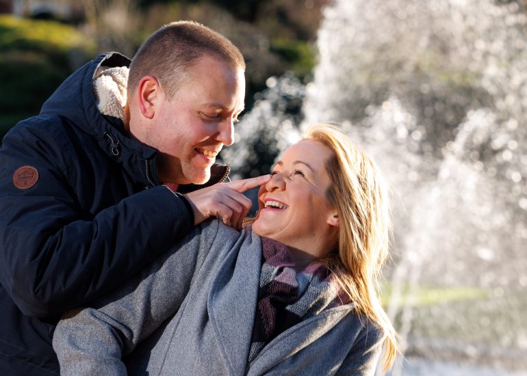 A Surrey Wedding Photographer captures a heartwarming moment between a man and woman during their engagement session at RHS Wiseley, as they share a tender hug in front of the fountain.