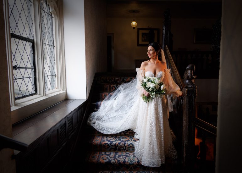 A bride in an elegant off-shoulder gown holding a bouquet, standing on a staircase beside a window in a dimly lit room at Eastwell Manor.