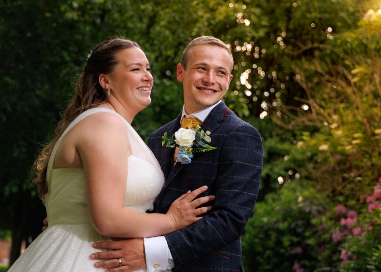 A bride in a white dress and a groom in a dark suit smile and embrace outdoors with greenery and flowers in the background, perfectly captured by Chilston Park wedding photography Kent.