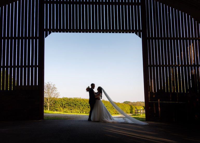 A bride and groom stand silhouetted against a bright outdoor backdrop inside the rustic charm of Hendall Manor Barns, with wooden slats and a gravel pathway completing the serene scene.
