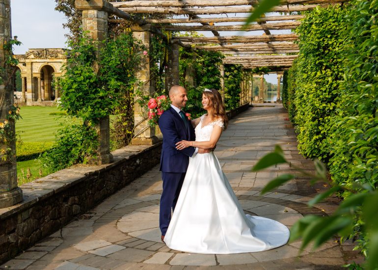 A bride in a white gown and a groom in a navy suit stand close together under a pergola surrounded by greenery. They are smiling and looking into each other's eyes, capturing the magic of their Hever Castle wedding photography.