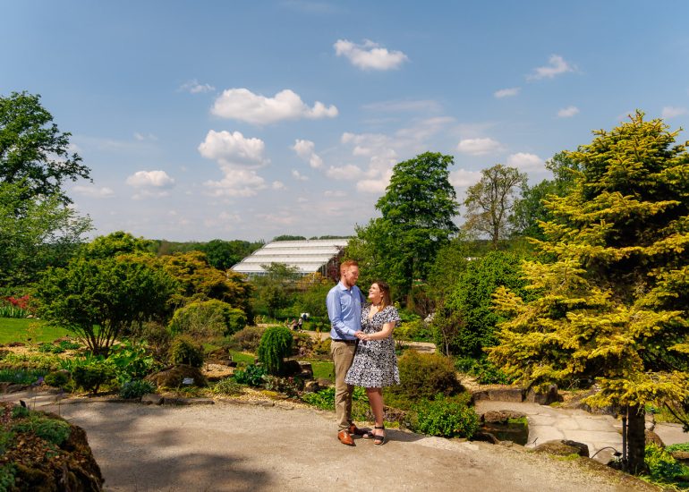 A couple stands together in a lush, scenic garden under a clear blue sky with white clouds. A greenhouse is visible in the background amidst the greenery. Captured by Sussex Wedding Photographer at RHS Wisley, this moment embodies romantic serenity.