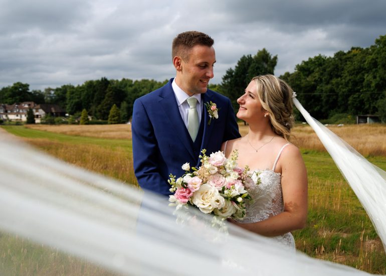 A bride and groom stand outdoors, smiling at each other; the bride holds a bouquet of flowers and her veil flows in the wind.