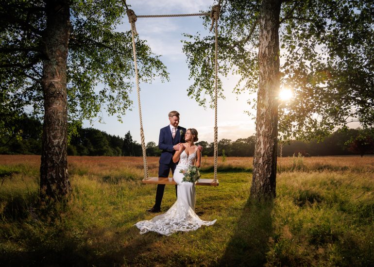 A bride in a white dress sits on a wooden swing, holding a bouquet, while the groom in a suit stands beside her. They are in a grassy field with trees and sunlight shining through, perfectly captured by Brookfield Barn Wedding Photography.
