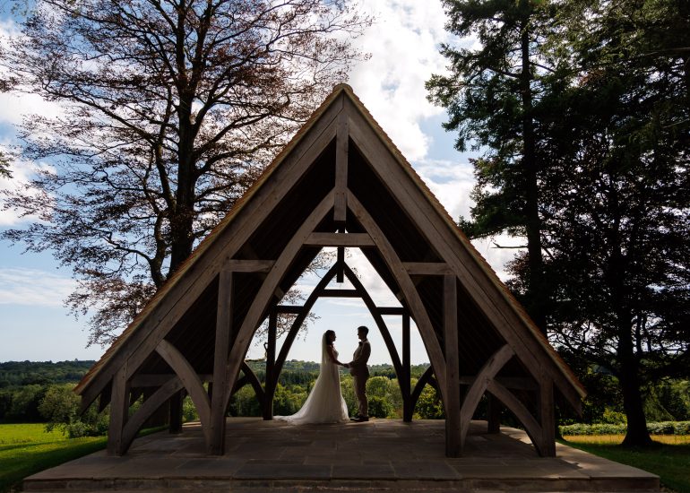 A couple stands facing each other under a wooden pavilion with a scenic green landscape and trees in the background, captured perfectly by Highley Manor Weddings Photographer.