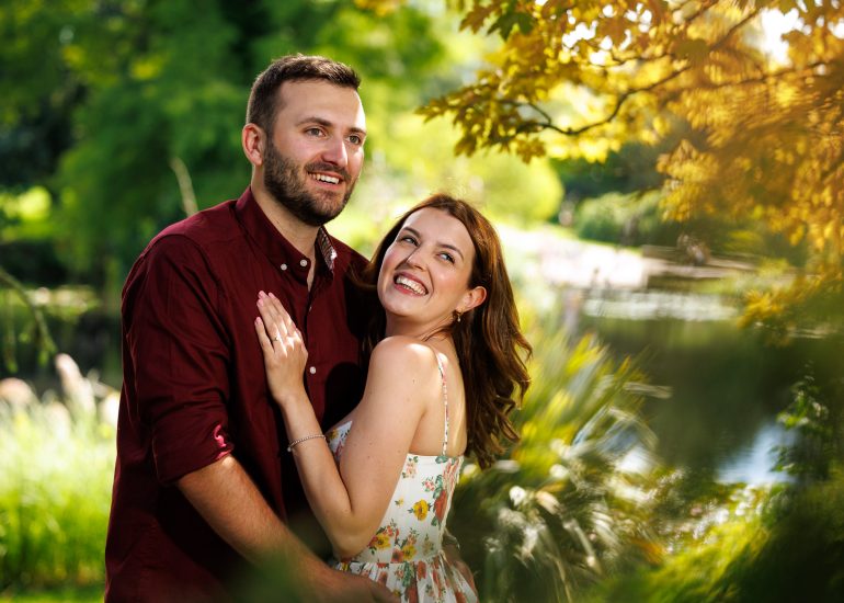 A couple stands outdoors in a leafy park at Wakehurst Place. The woman is smiling and looking up at the man, who is looking off into the distance. They appear happy and content, capturing a special moment that highlights the beauty of wedding photography.