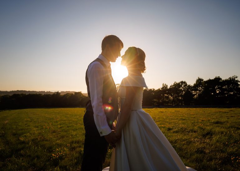 A couple holds hands and faces each other in an open field at sunset, with the sun positioned between them, creating a silhouette effect—a moment beautifully captured by Sussex Barn Wedding Photography.