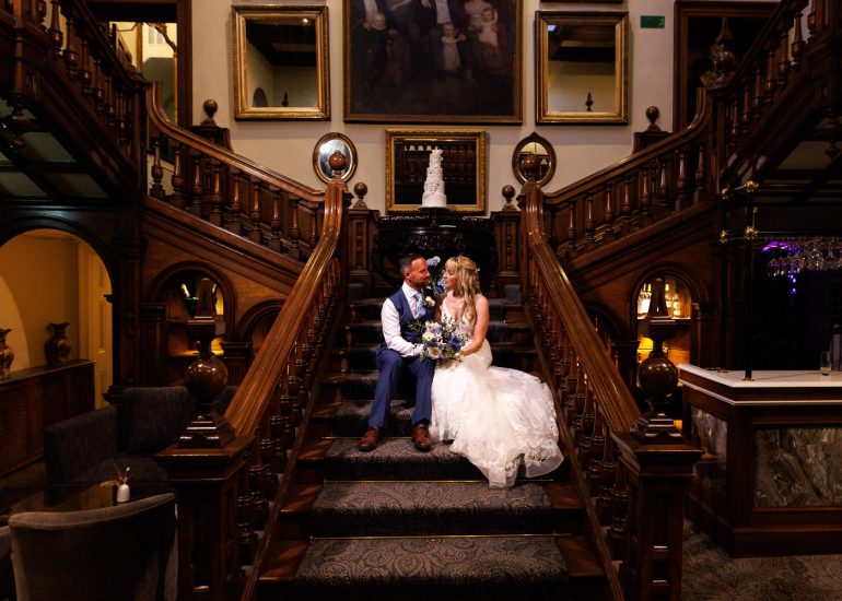 A bride and groom sit together on an ornate wooden staircase inside a beautifully decorated venue at Chilston Park Hotel, holding a bouquet.