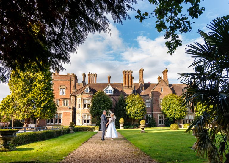 A couple in wedding attire stands on a pathway at the picturesque Alexander House Hotel, framed by a historic brick building surrounded by trees and a well-kept garden.