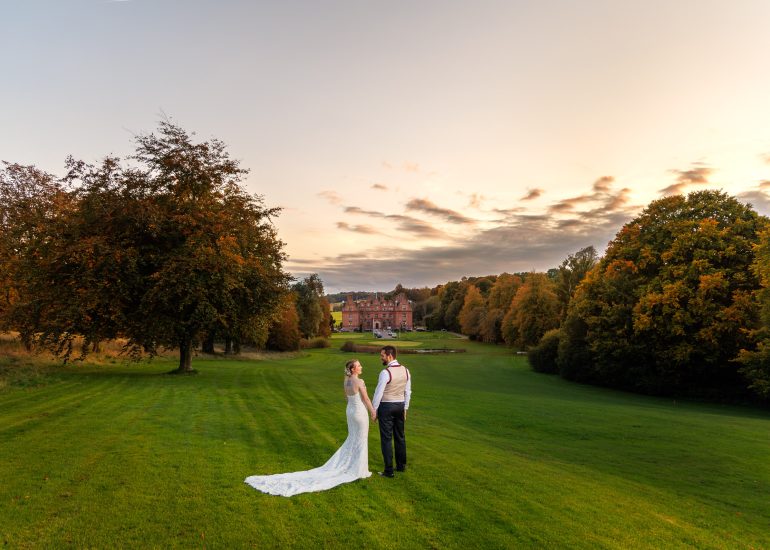 A couple in formal attire stands on a grassy field with the majestic Kent Hotel in the background, capturing the enchanting essence of sunset weddings.