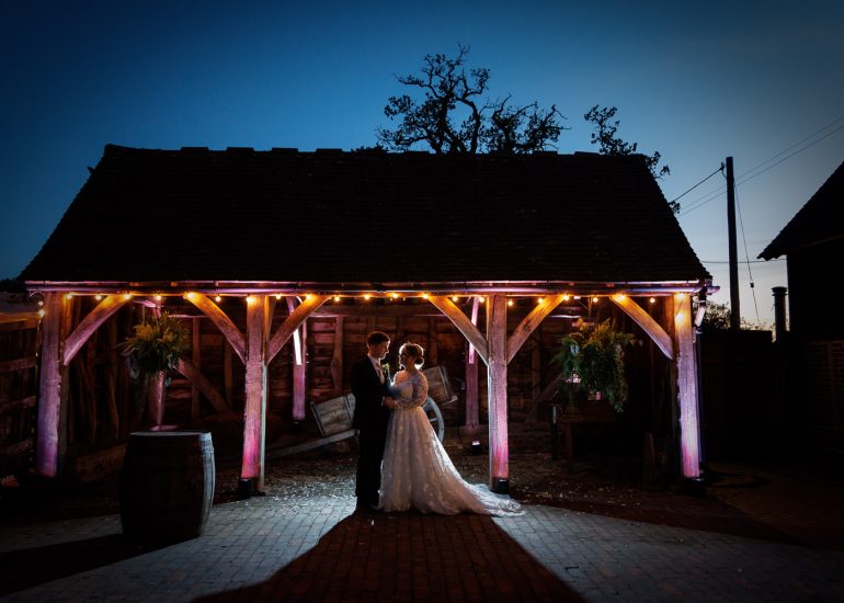 A bride and groom stand facing each other under a wooden shelter with string lights at dusk.
