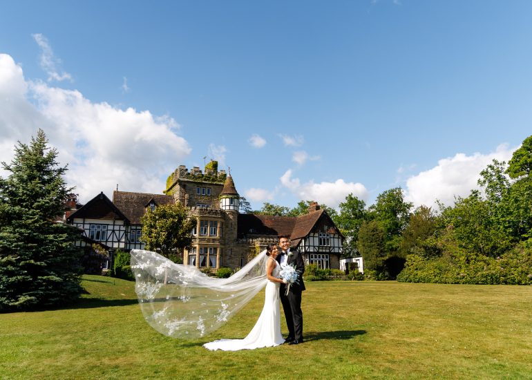 A bride and groom stand together on a large grassy lawn in front of a historic manor at The Ravenswood Weddings, with the bride’s veil flowing in the breeze under a clear blue sky.