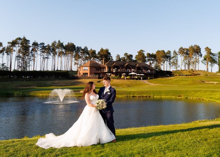 A bride and groom stand together by a pond with a fountain, on a grassy area in front of a large building surrounded by trees under a clear blue sky—an idyllic setting for Westerham Golf Club Weddings.