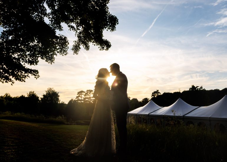 Silhouetted couple stands close together at sunset near white event tents at Swarling Manor Weddings, with trees and a clear sky in the background.
