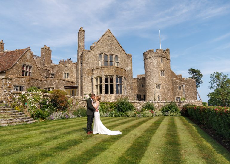 A bride and groom stand embracing on a striped lawn in front of a large historic stone building under a clear sky, capturing the timeless romance of Lympne Castle weddings.