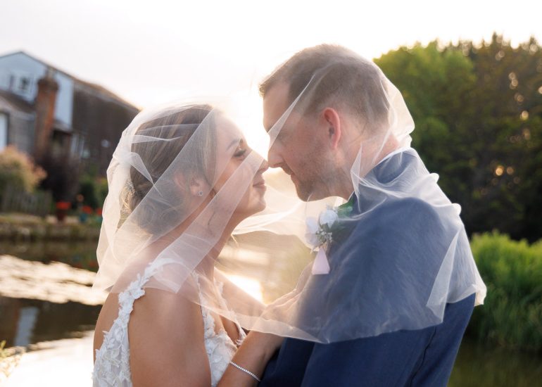 A bride and groom stand outdoors at Coltsford Mill Wedding Venue in Surrey, facing each other closely under a veil, smiling, with greenery and water in the background.