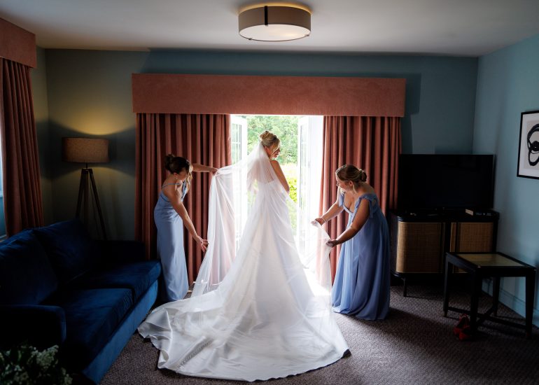 A bride in a white gown stands near an open door at Lythe Hill Hotel Spa Wedding Venue in Surrey, as two bridesmaids in light blue dresses adjust her train in a cozy living room, capturing a beautiful photography moment.