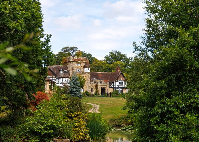 A large stone and timber house with gabled roofs and a tower sits in a landscaped garden, surrounded by tall green trees and a pond in front—an ideal setting for The Ravenswood in West Sussex wedding photographer to capture magical moments.