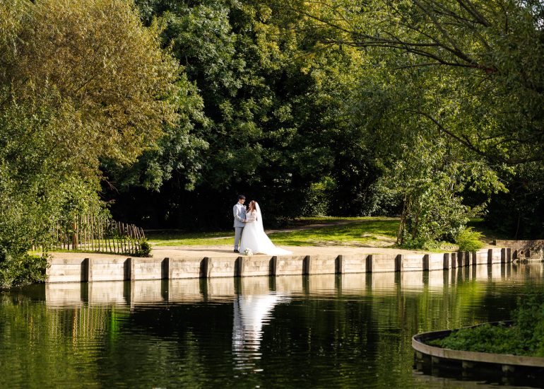 A couple in wedding attire stands together on a path by a pond, surrounded by trees and greenery, their reflection visible in the water—capturing the essence of Kent wedding photography.