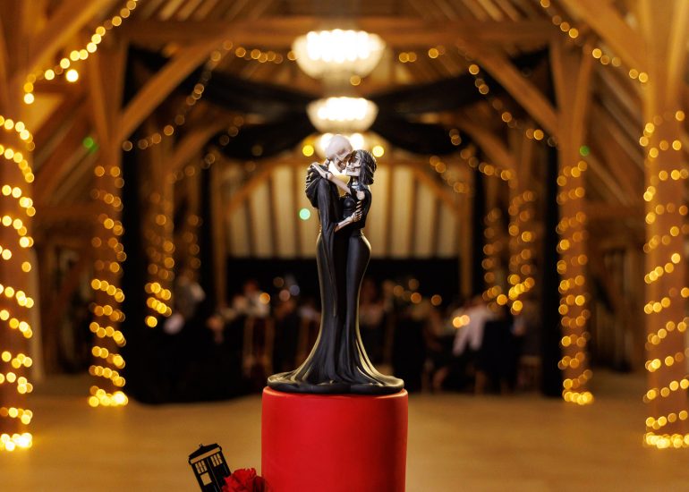 A cake topper of two figures embracing, standing on a red cake, with a decorated wooden hall and string lights in the background.